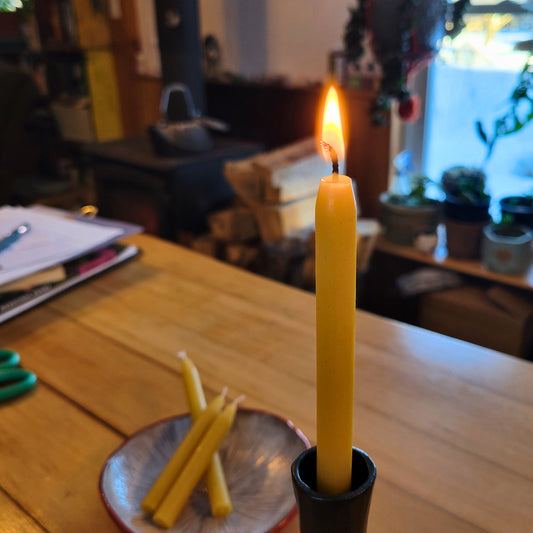 Tall beeswax mini-taper burns in a taper holder while unlit candles lie on a mushroom plate in the background.