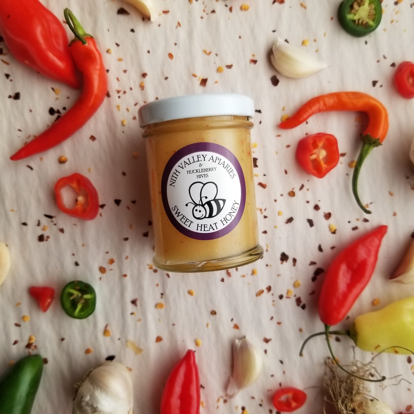Sweet Heat Honey 90g Jar displayed with garlic and hot peppers artfully arranged in the background.