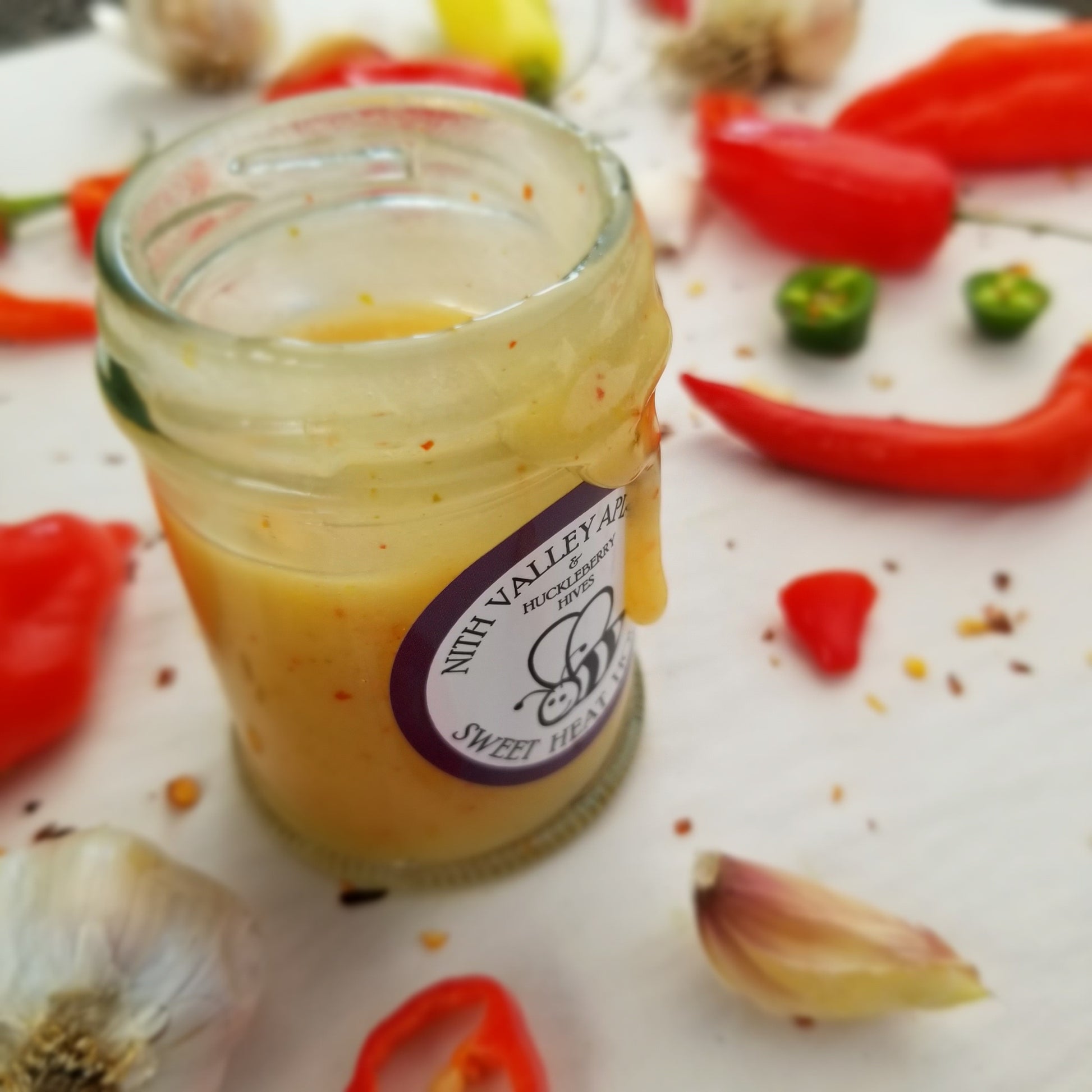 Sweet Heat Honey 90g Jar opened with a honey drizzle. Garlic and hot peppers artfully arranged in the background.