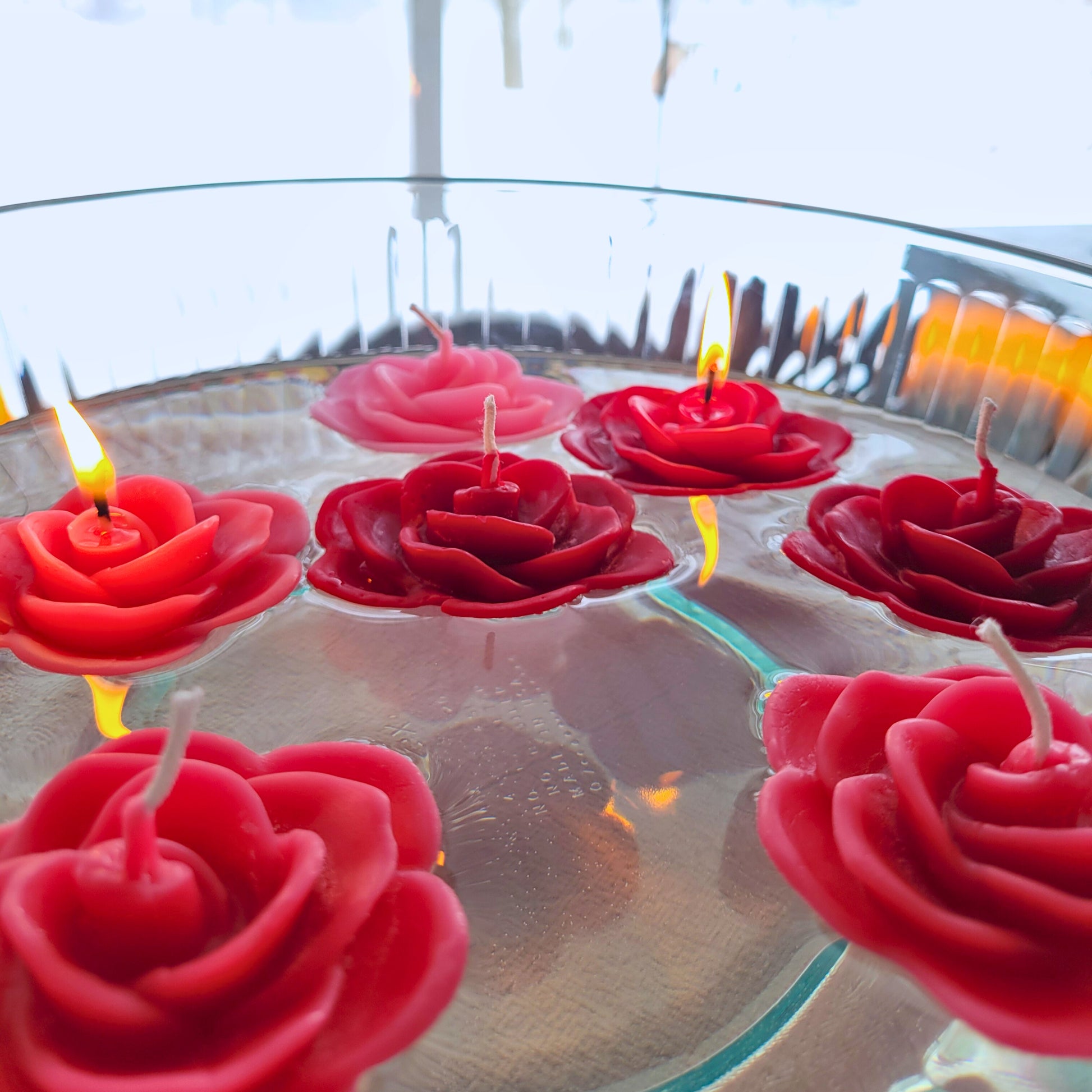 Red and pink rose-shaped candles float in a bowl of water with a blurred outdoor background