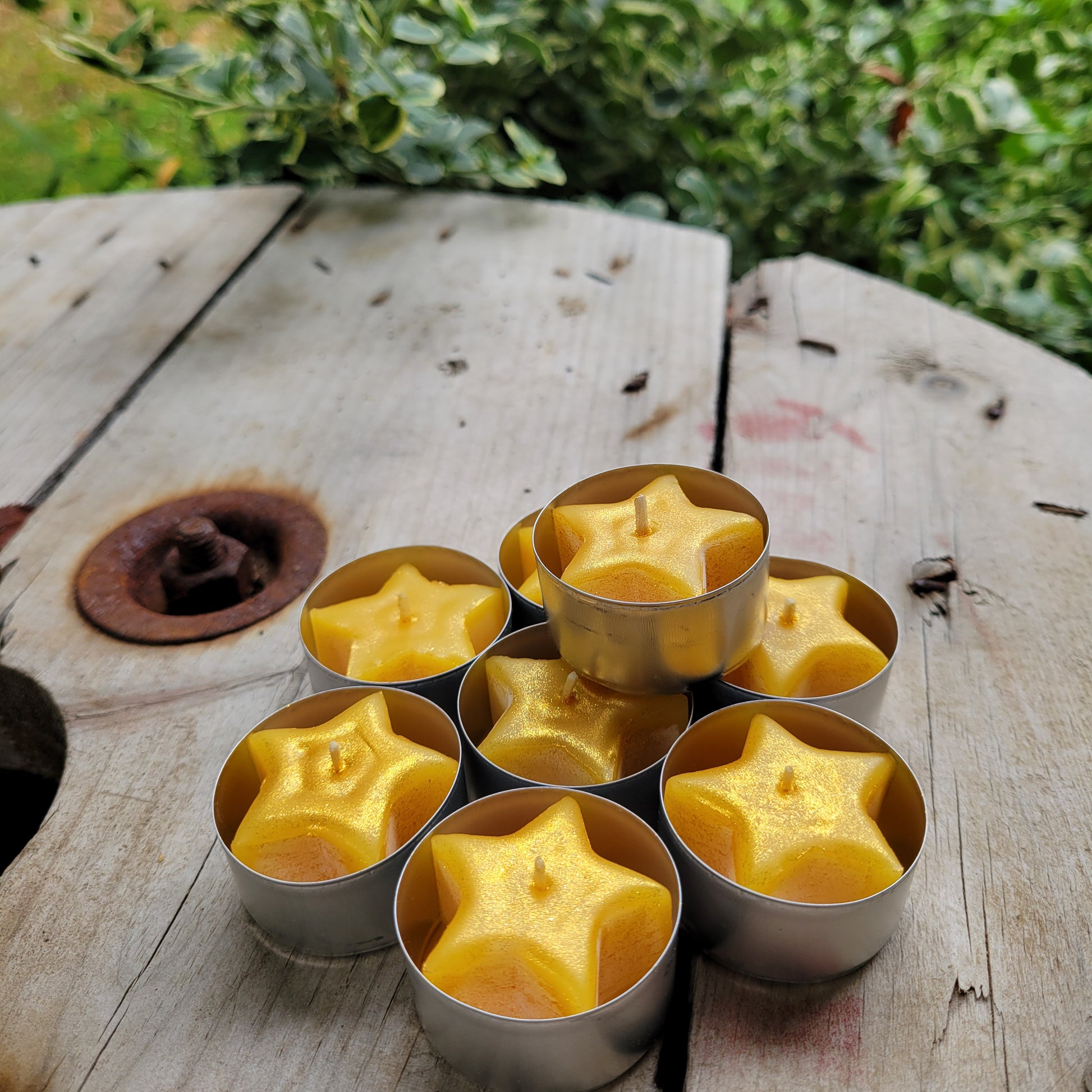 Star-shaped sparkly yellow beeswax candles in metal tealight cups on a wooden surface with greenery in the background